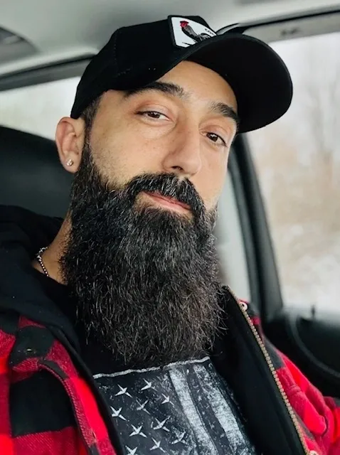 A man with long black beard and cap sitting in the back of a car.